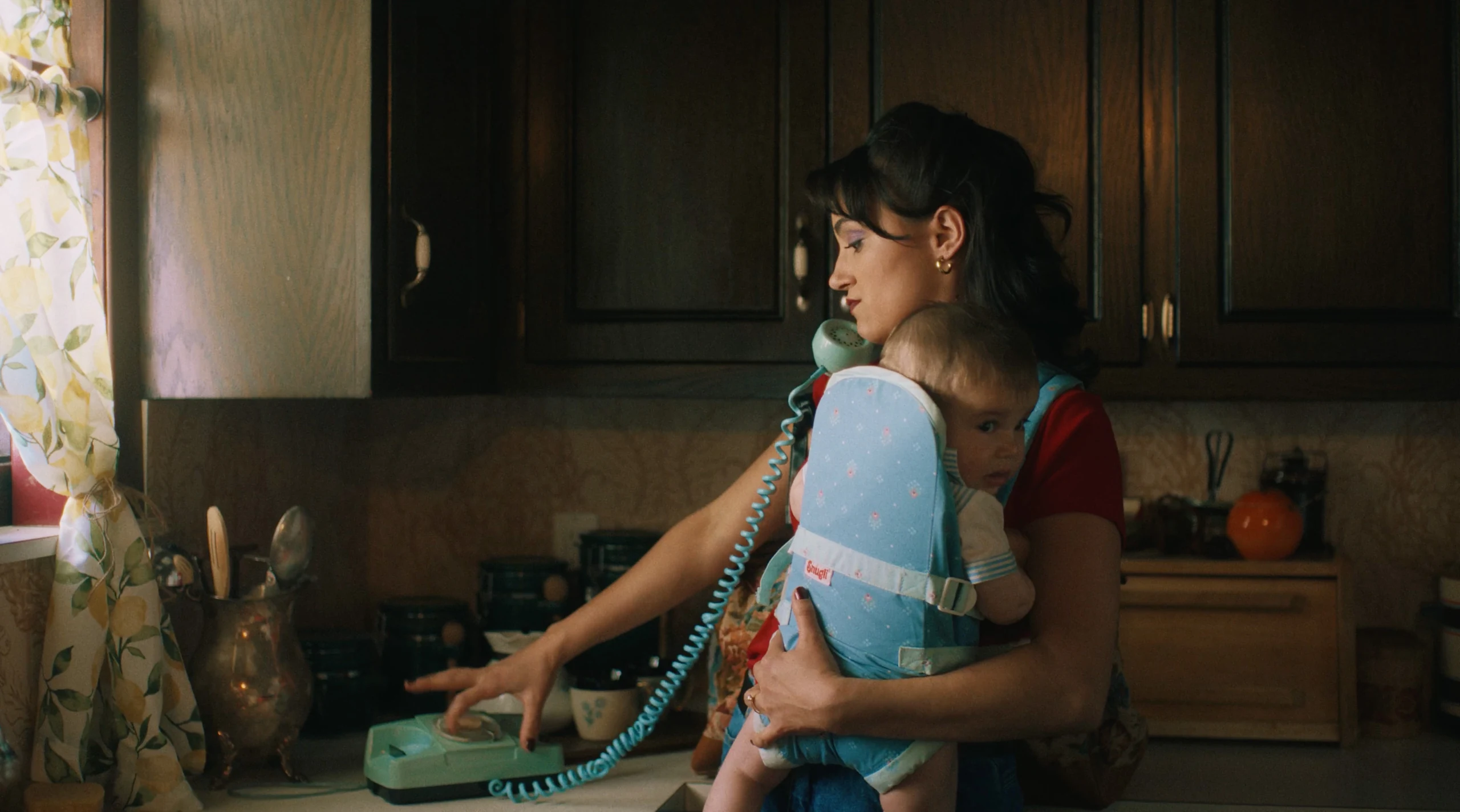 A woman in a red T-shirt holds a baby in a blue carrier while dialing a mint rotary phone in a wood-cabinet kitchen
