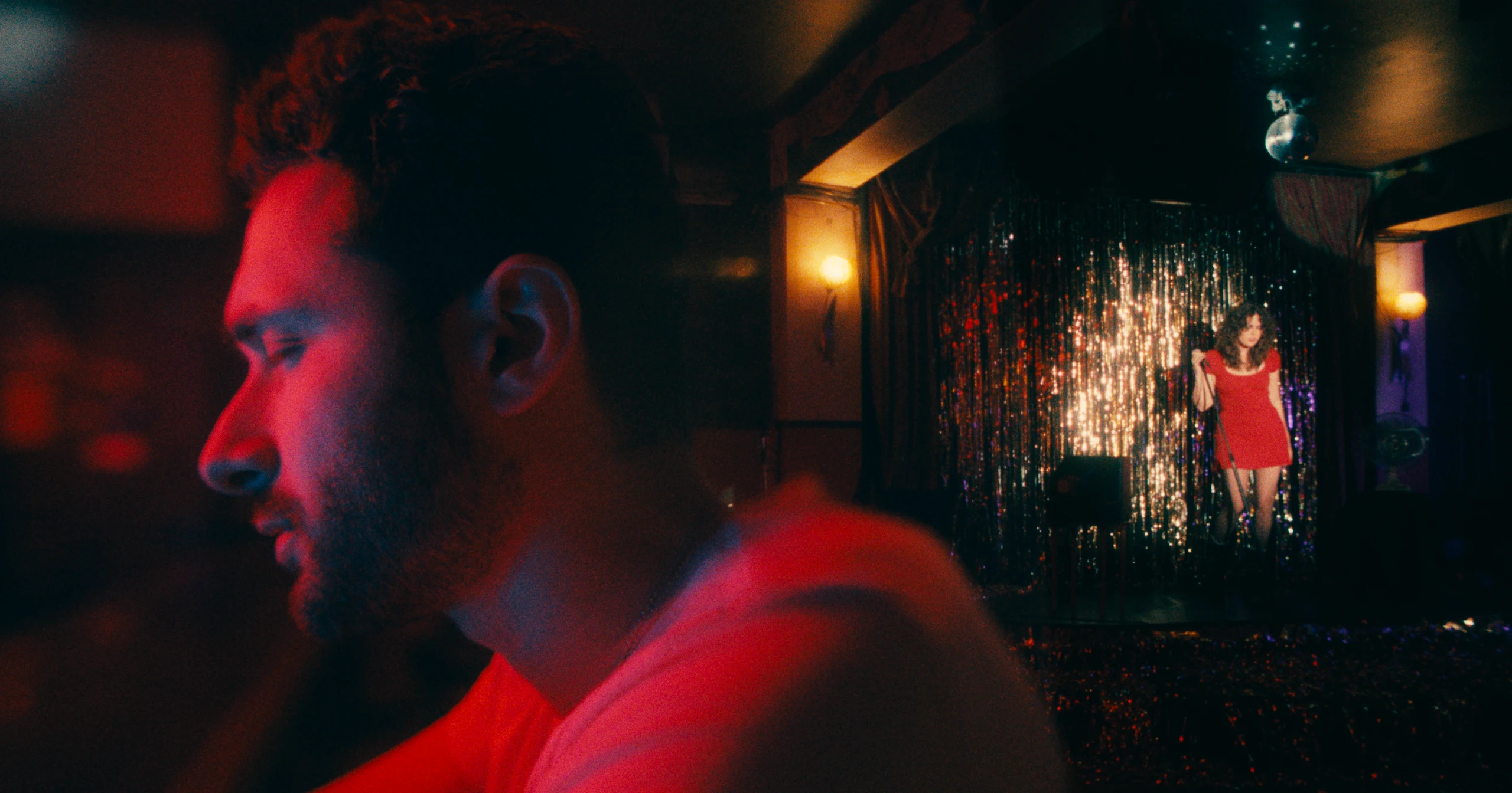 A bearded man lit red in the foreground, with a woman in a red dress on a tinsel-curtained stage behind