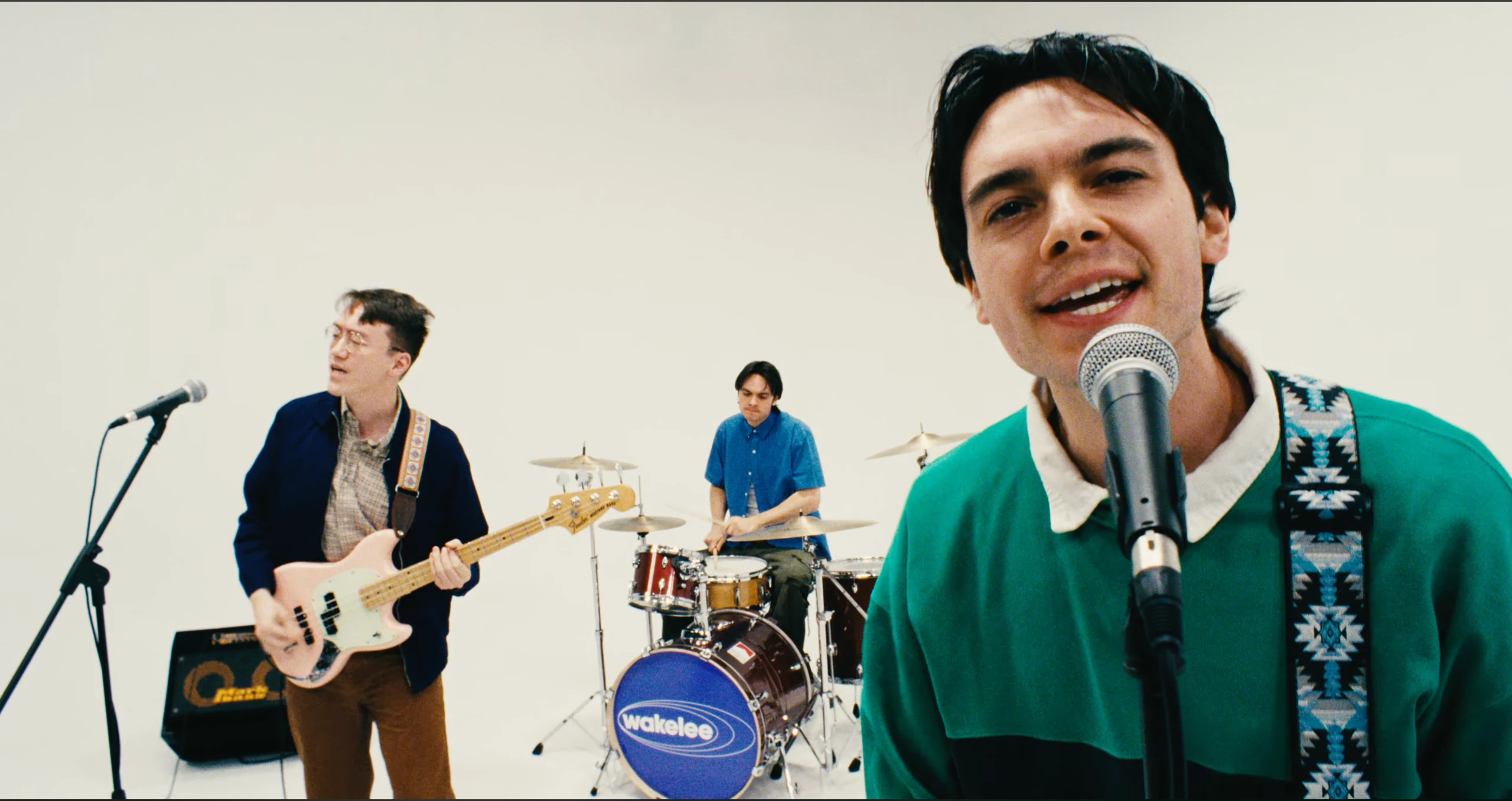 A singer wearing green leans toward a microphone, with a bassist and drummer performing behind him in a white studio.